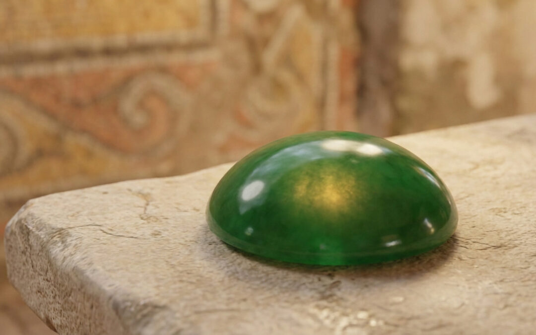 A magnificent, smooth emerald jade cabochon, glowing golden from within, on an ancient beige stone, with blurred mosaic background.