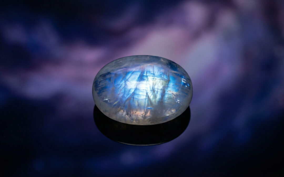A radiant, iridescent moonstone with blue, white, and silver flashes glows on a dark, reflective surface, against a mystical indigo and lavender background.