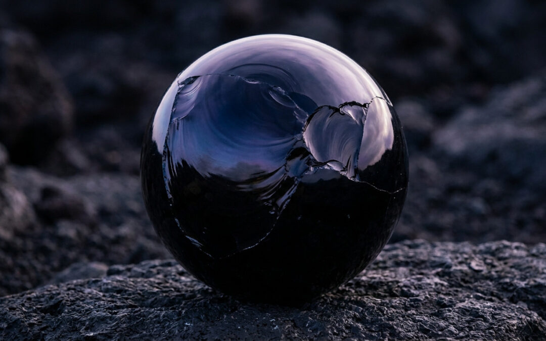 A high-resolution, polished black obsidian sphere with indigo and violet reflections, resting on dark volcanic rock. Shows conchoidal fractures and dramatic lighting.